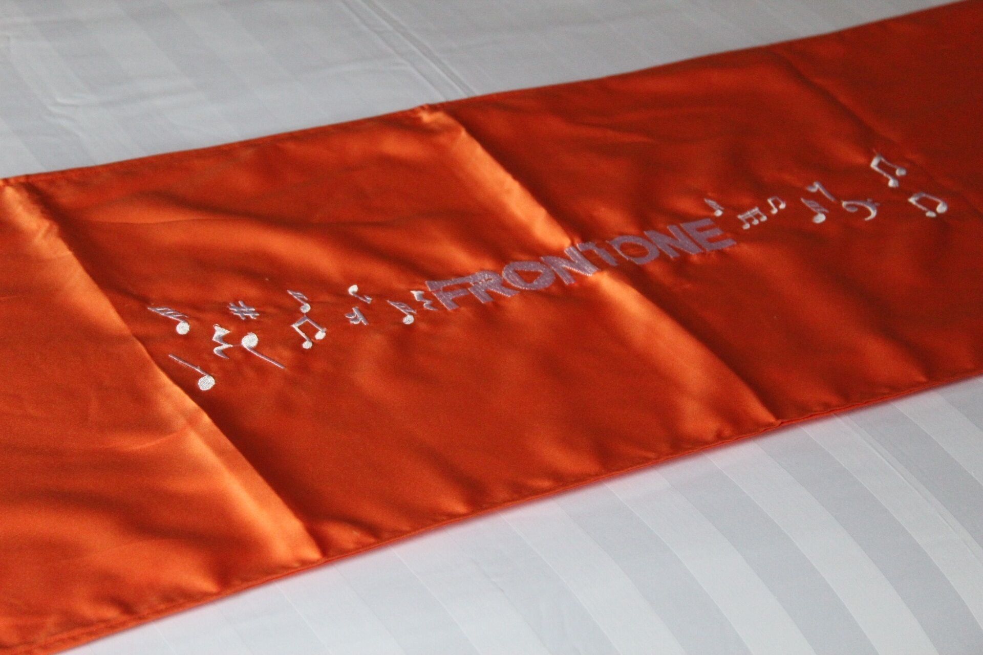 Una cama con sábana blanca y almohada naranja con notas musicales bordadas y la palabra "FRONTONE".