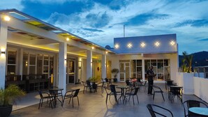 Ein Terrassenbereich im Freien mit Tischen und Stühlen, eine überdachte Sitzgelegenheit und ein Gebäudeeingang mit Glasüren.