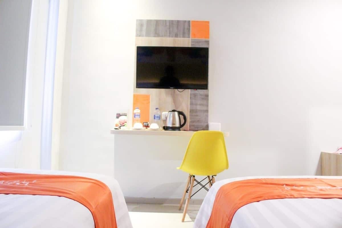 Ein Hotelzimmer mit zwei Betten, einer kleinen Küchenzeile, einem Flachbildfernseher und einem leuchtend gelben Sessel.