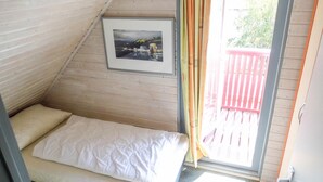 Ett litet rum med en enkel säng, ett sluttande tak, en inramad tavla på väggen och utsikt över ett rött trädäck genom en glasdörr.