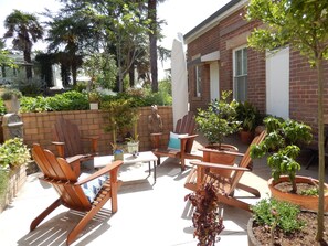 Un patio con sillas y mesas de madera, rodeado de plantas en macetas y un muro de ladrillo.