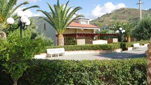 A two-story house with a red roof, surrounded by lush greenery and palm trees. - Residence Fontana Barone (Cefalù)