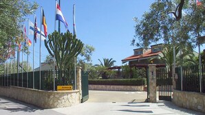 A gated entrance with multiple flags, a stone wall, and a sign indicating 'Residence Fontana Barone'. - Residence Fontana Barone (Cefalù)