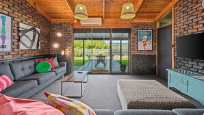 A cozy living room with a sofa, a coffee table, and a patterned ottoman. There is a television on a stand and a fireplace. The room has wooden ceilings and brick walls. - Black Pearl - Aldinga Beach - C21 SouthCoast Holidays (Aldinga Beach)