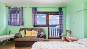 Una stanza con un divano marrone, un letto, un tavolino con una lampada e vista sul mare attraverso una finestra con tende viola.