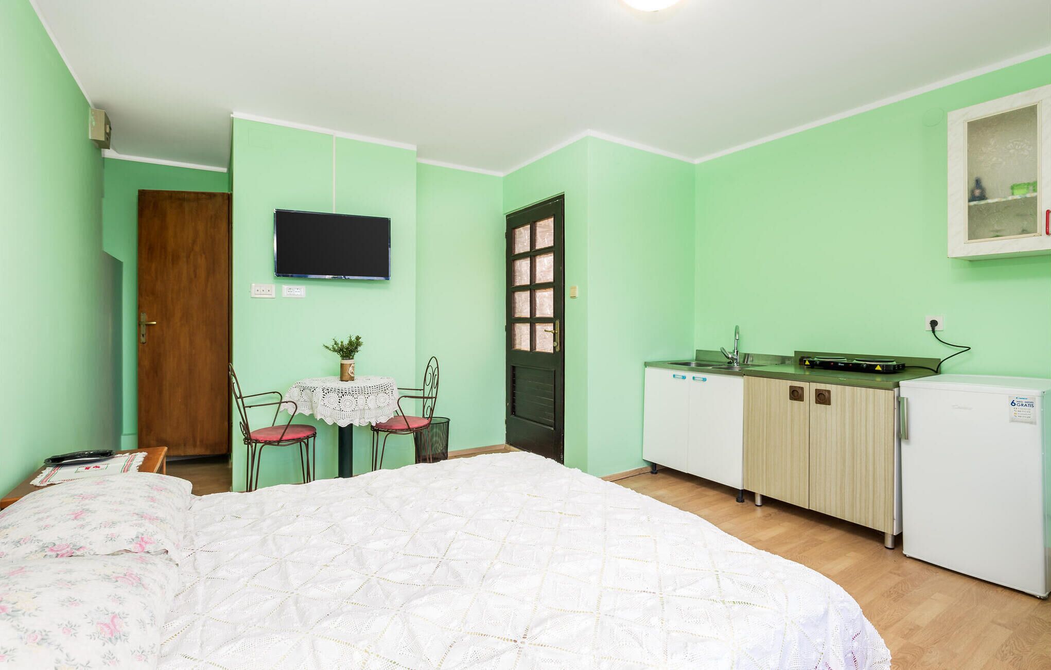 A room with a bed, a small table with chairs, a kitchenette, and a television on the wall.