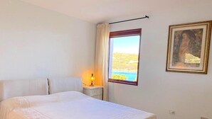 A bedroom with a large bed, a nightstand with a lamp, a window with a view of the sea, and a framed picture on the wall. - Sea Front Bonbonnière - Porto Cervo Costa Smeralda (Porto Cervo)
