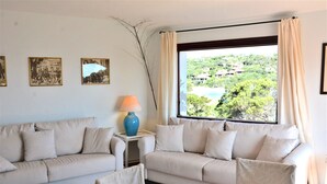 A room with two beige sofas, a small table with a lamp, and a view of a green landscape through a large window. - Sea Front Bonbonnière - Porto Cervo Costa Smeralda (Porto Cervo)