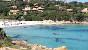 A coastal area with clear blue waters, a sandy beach, and houses on a hillside. - Sea Front Bonbonnière - Porto Cervo Costa Smeralda (Porto Cervo)
