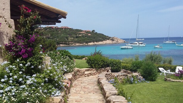 A stone pathway leading to a coastal area with boats on the water, greenery, and a building with a roof. - Sea Front Bonbonnière - Porto Cervo Costa Smeralda (Porto Cervo)
