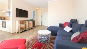 A modern living room with a blue sofa, red cushions, a round coffee table, and a kitchen area with white cabinets and a TV.