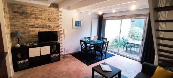 A living room with a brick wall, a flat-screen TV, a dining table with chairs, and a sliding glass door leading to an outdoor area.