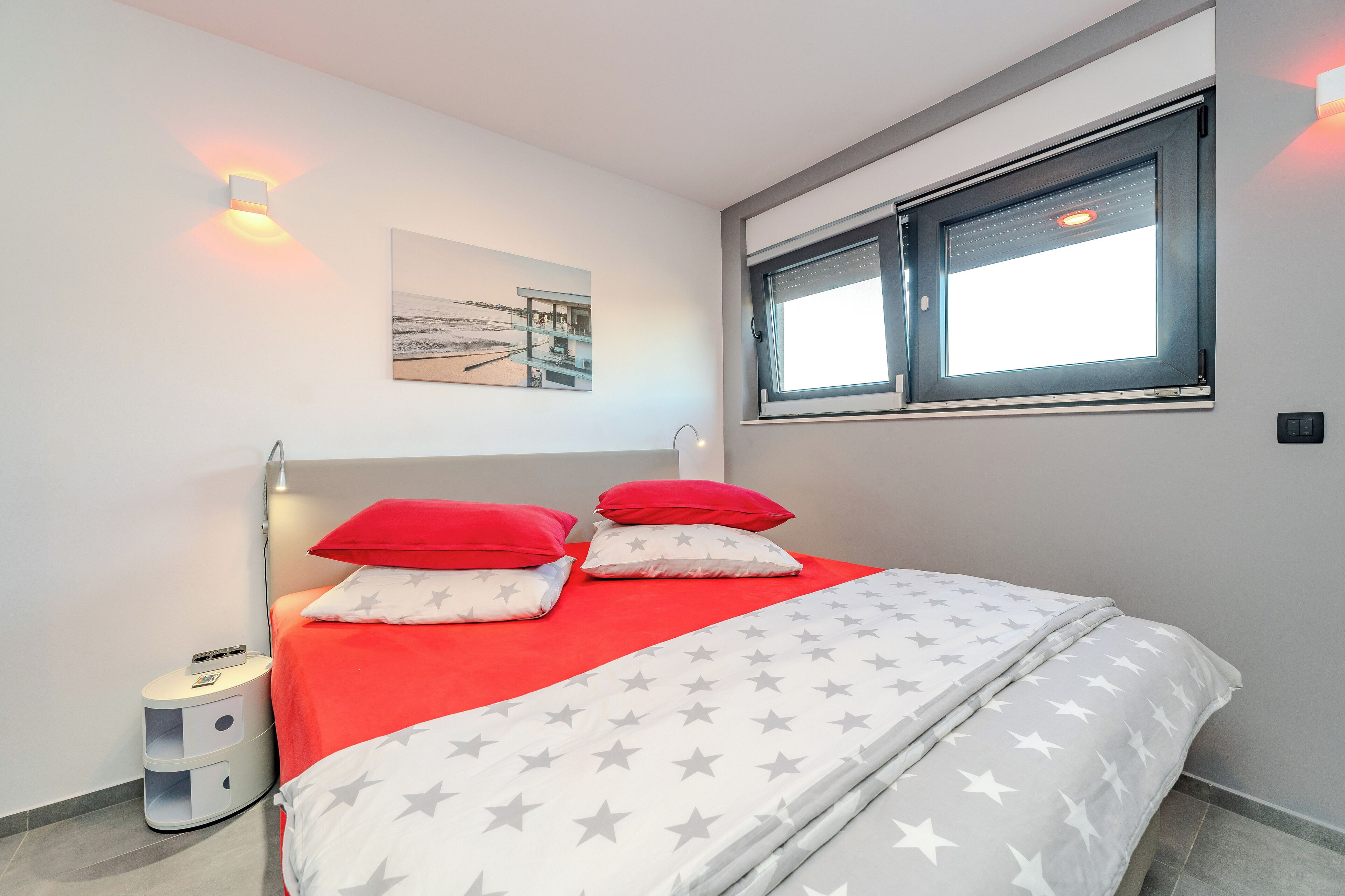 Een moderne slaapkamer met een bed, rode en witte kussens, een deken met sterrenprint, een nachtkastje met lamp en een raam met daglicht.