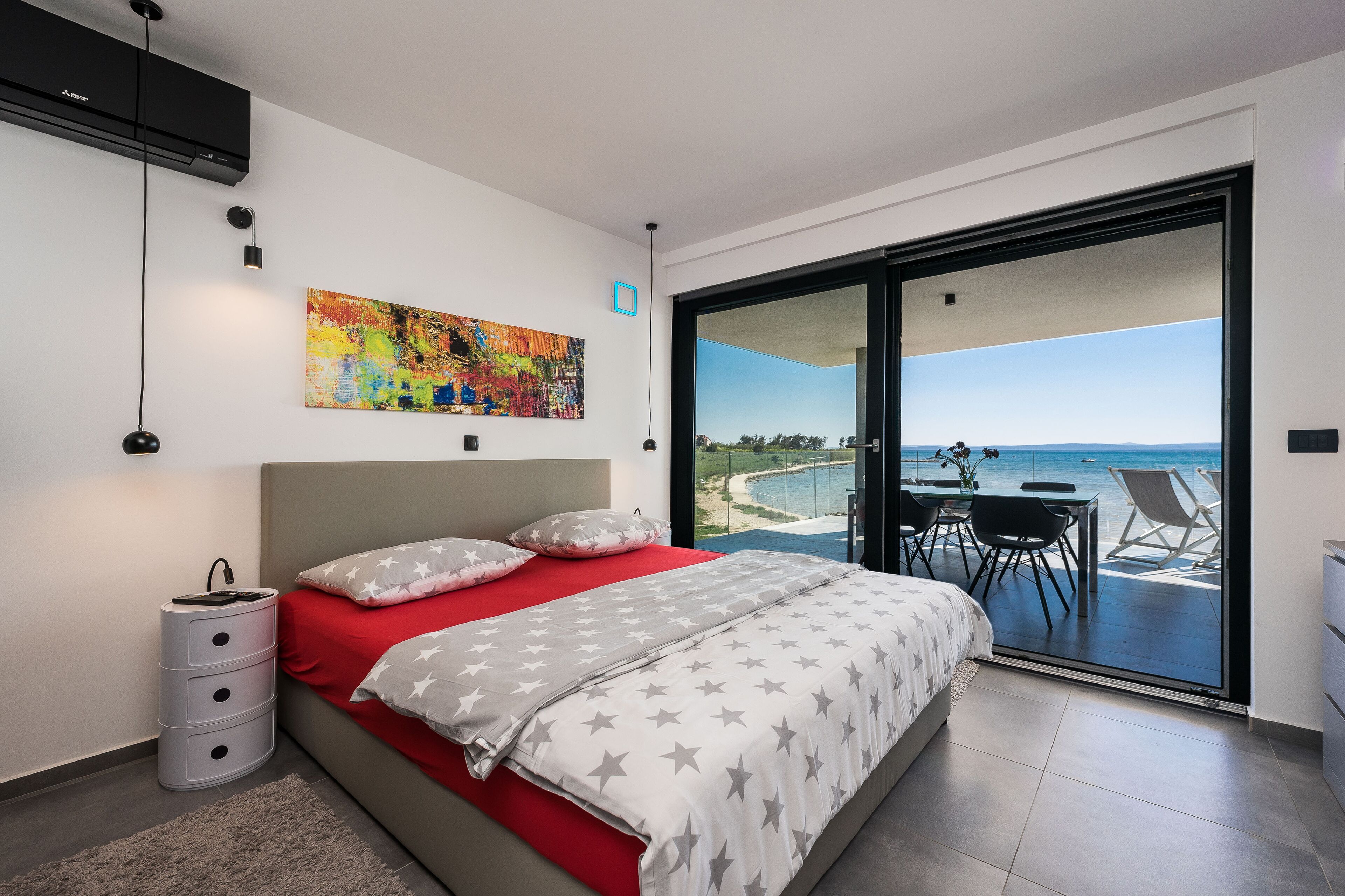 Ein modernes Schlafzimmer mit einem Bett, einem farbenfrohen abstrakten Gemälde, einem Schreibtisch mit Stühlen und Meerblick durch eine Schiebeglastür.