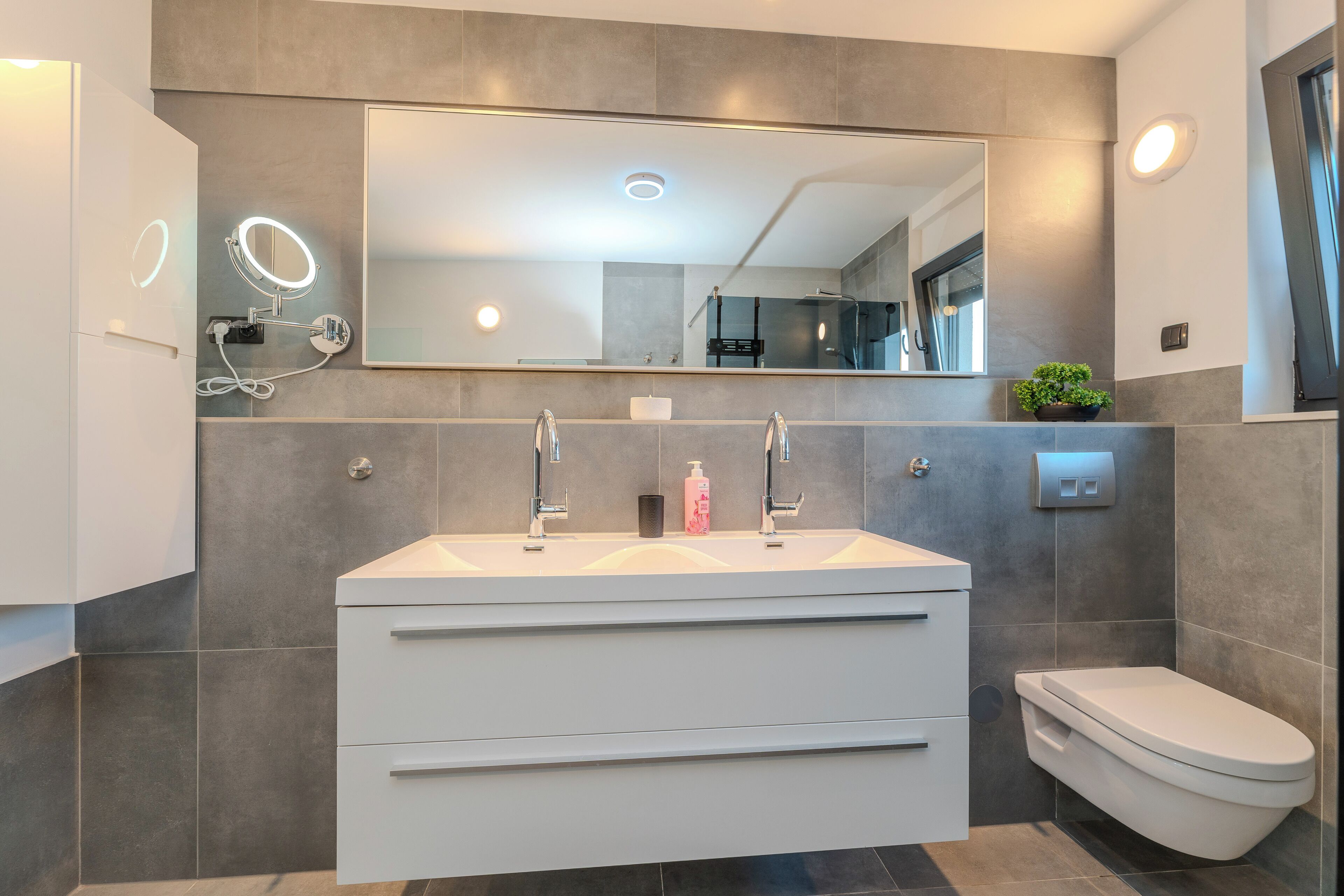 A modern bathroom with a large mirror, two sinks, a toilet, and a vanity with drawers.