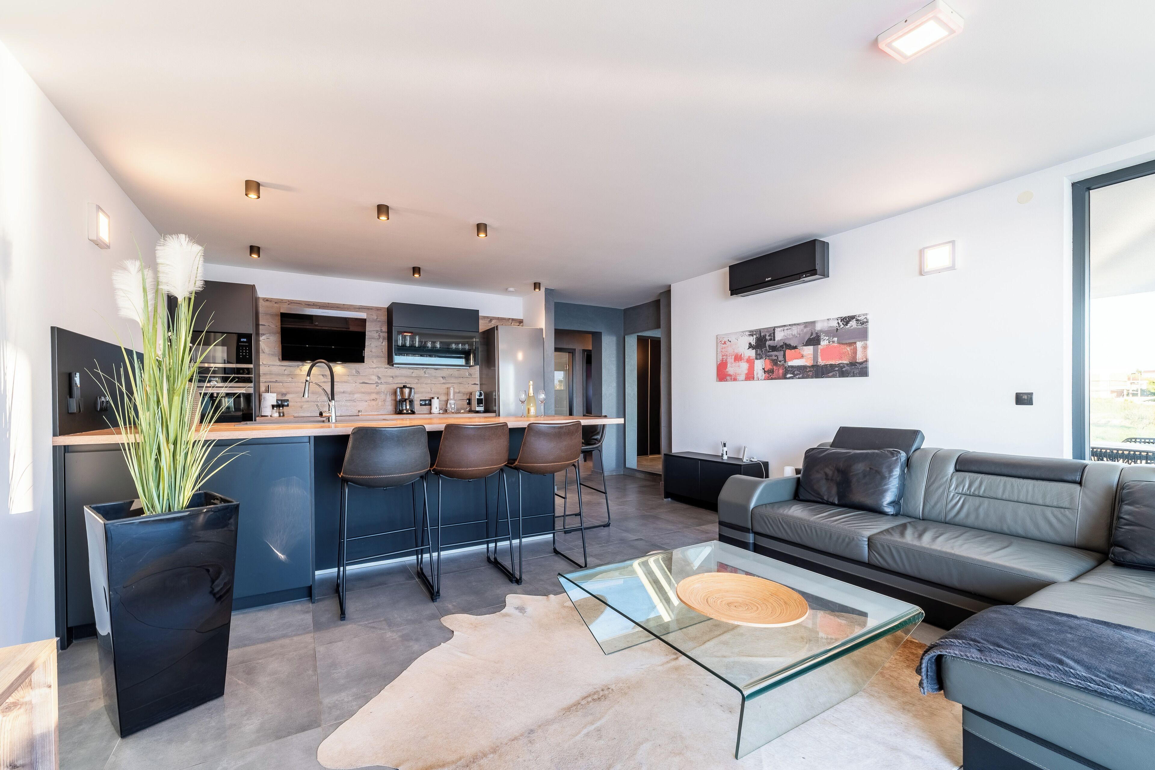 A modern living room with a glass coffee table, leather sofa, and a kitchen area with bar stools.