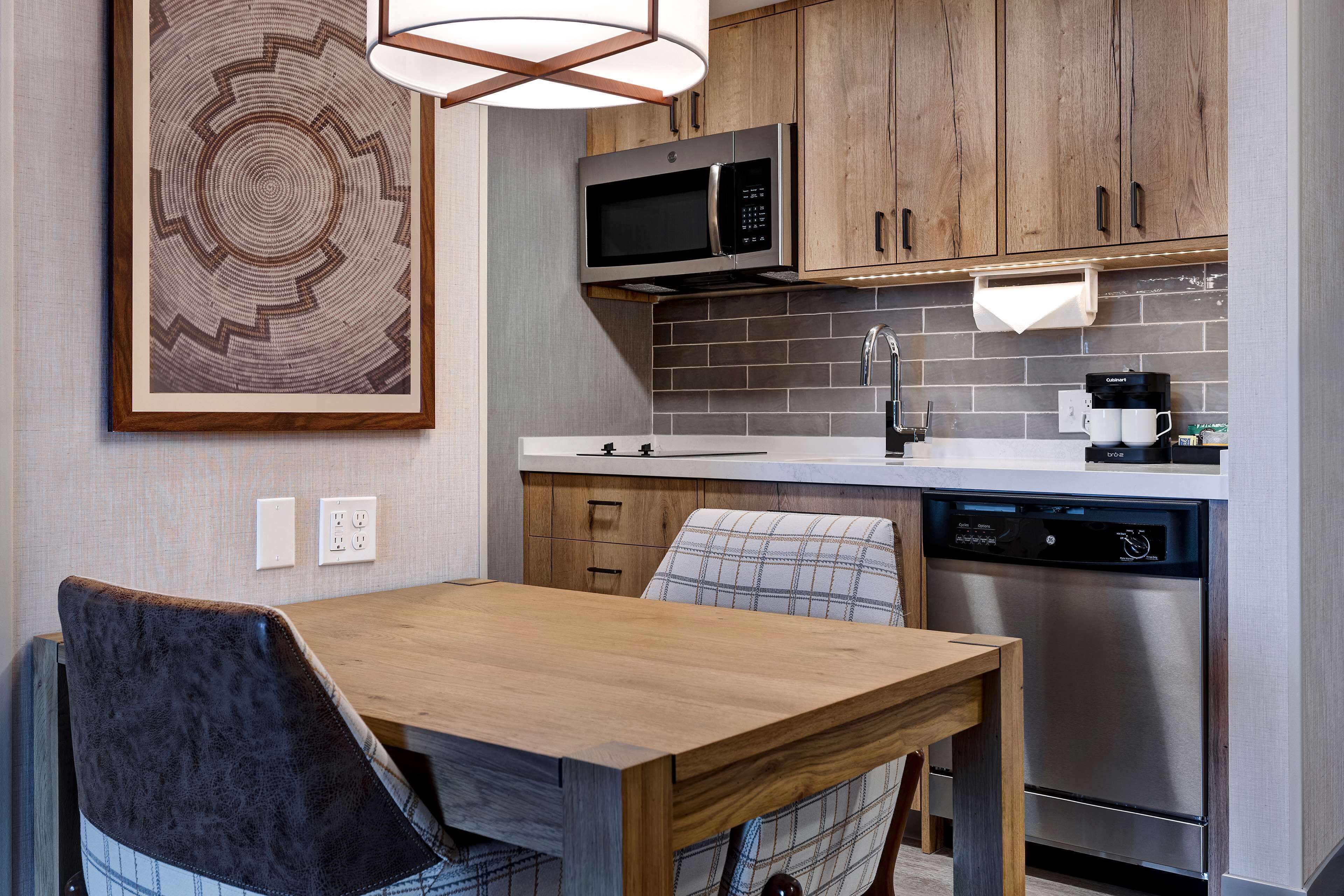 A modern kitchen with a stainless steel dishwasher, a microwave, and a coffee maker. There is a wooden dining table with two chairs.
