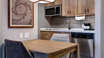 A modern kitchen with a stainless steel dishwasher, a microwave, and a coffee maker. There is a wooden dining table with two chairs.