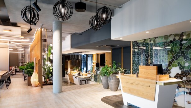 Een moderne receptiebalie met een houten toonbank, grote potplanten en een mozaïekwand.