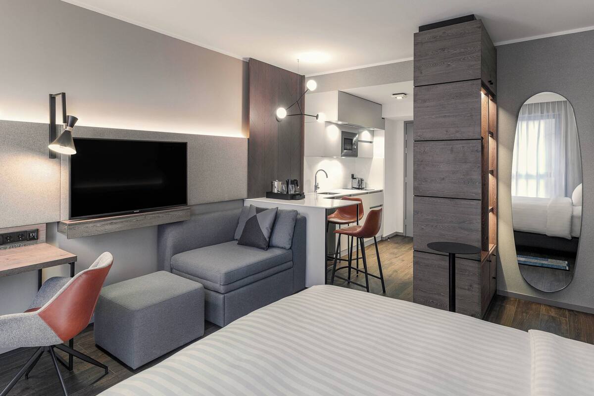 A modern hotel room with a large bed, a desk, a sofa, a TV, a kitchen area, and a bathroom with a mirror.