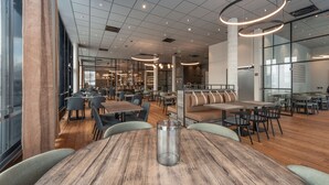 A modern restaurant with wooden tables and chairs, a glass partition, and a high ceiling with circular light fixtures.
