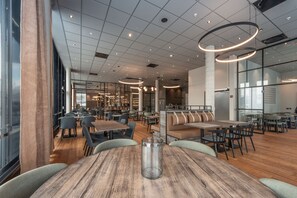 A modern restaurant with wooden tables and chairs, a glass partition, and a high ceiling with circular light fixtures.