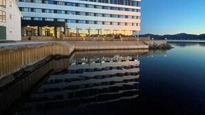 A modern hotel with a reflective pool in front, a wooden deck, and a view of the water and mountains in the background.