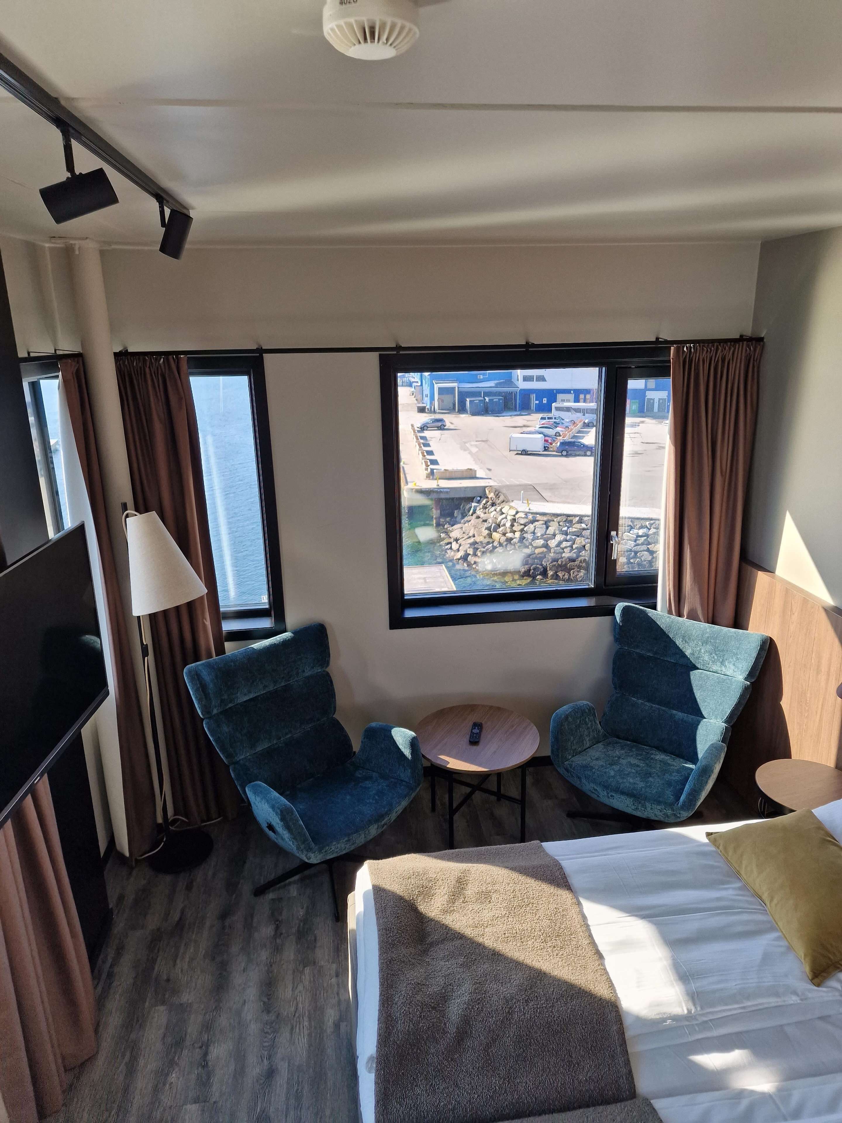 Habitación de hotel con cama, dos sillones, una mesita, televisor y ventana con vista a un estacionamiento y edificios.