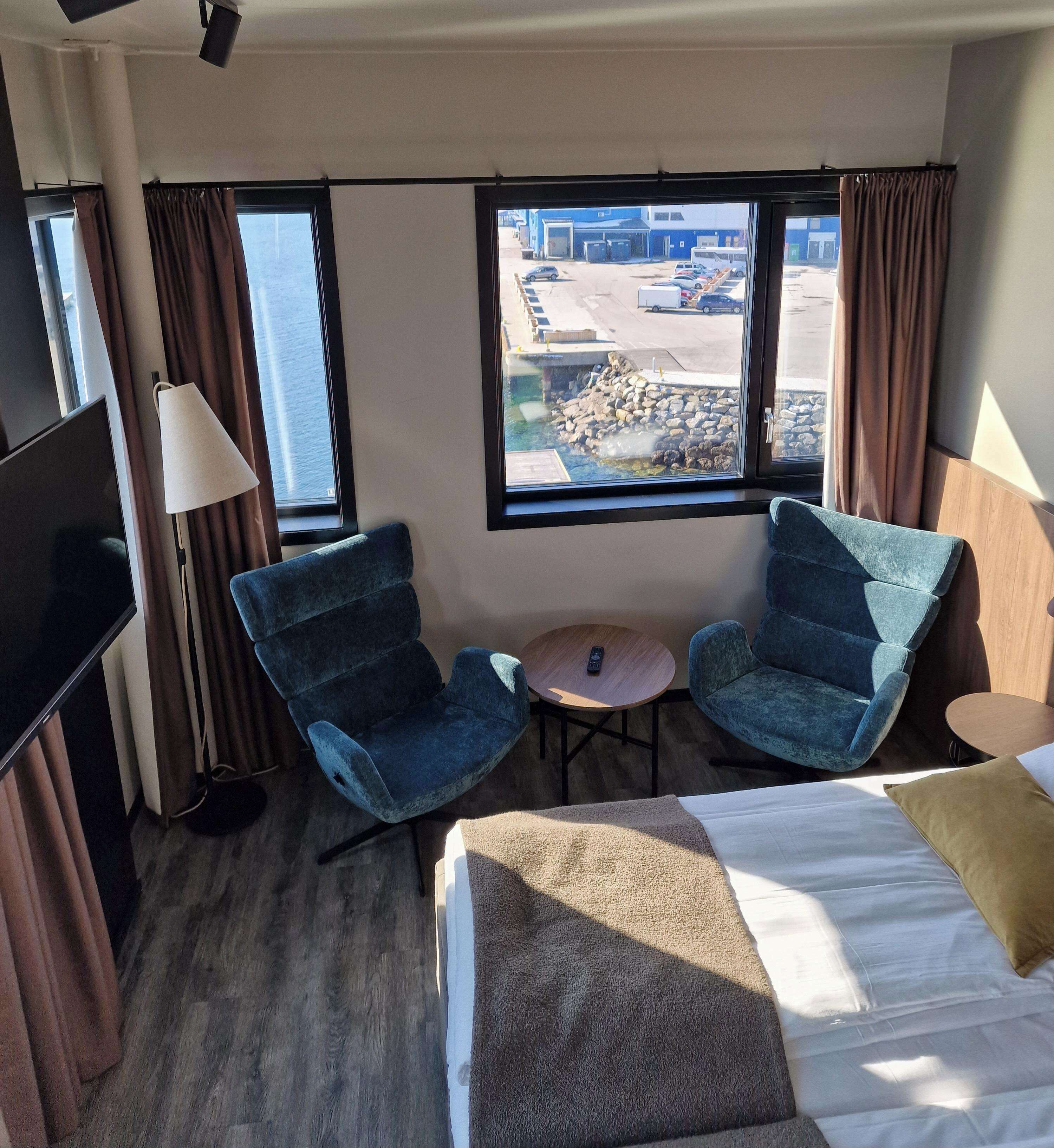Une chambre d’hôtel avec un lit, deux fauteuils bleus, une petite table ronde, un téléviseur et une fenêtre donnant sur un parking.