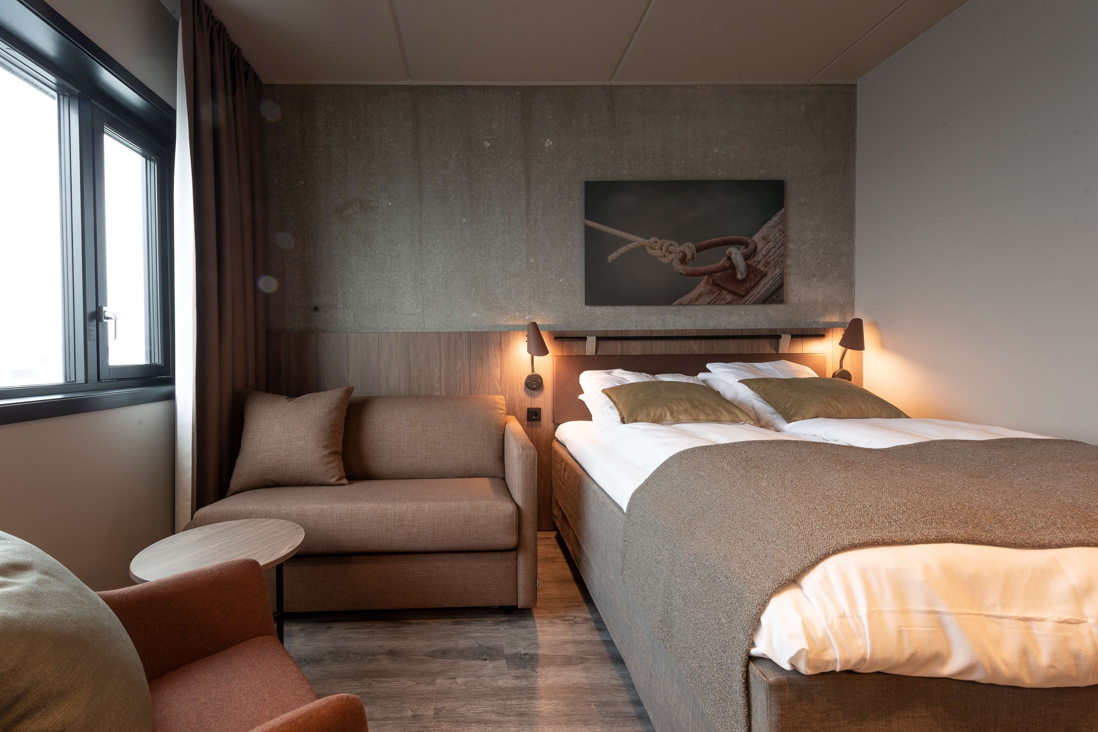 A modern hotel room with a bed, a sofa, a small table, and a painting on the wall.