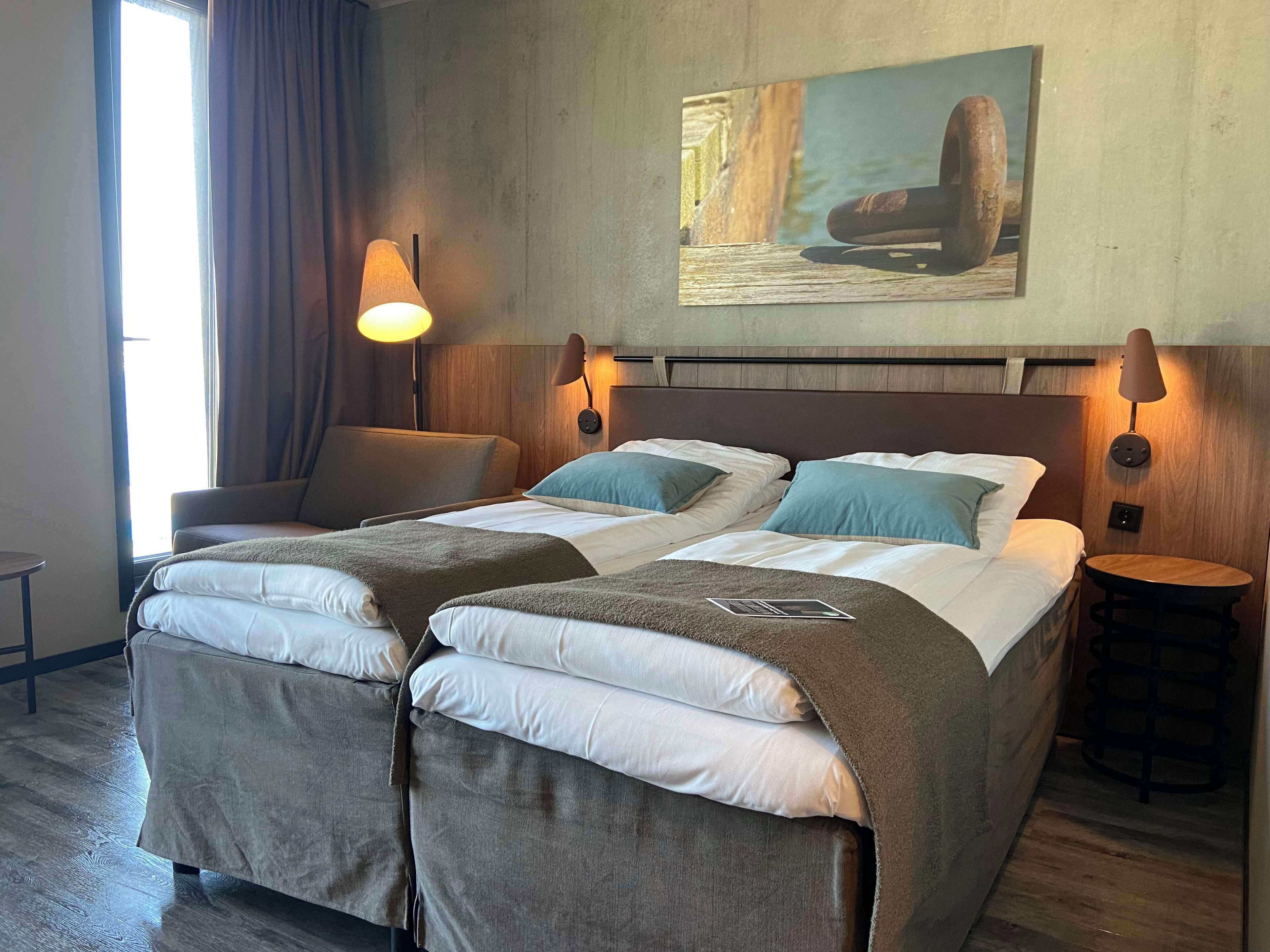 A double bed with two pillows, a bedside table, a lamp, and a chair in a room with a wooden headboard and a painting on the wall.