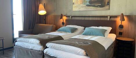 A double bed with two pillows, a bedside table, a lamp, and a chair in a room with a wooden headboard and a painting on the wall.