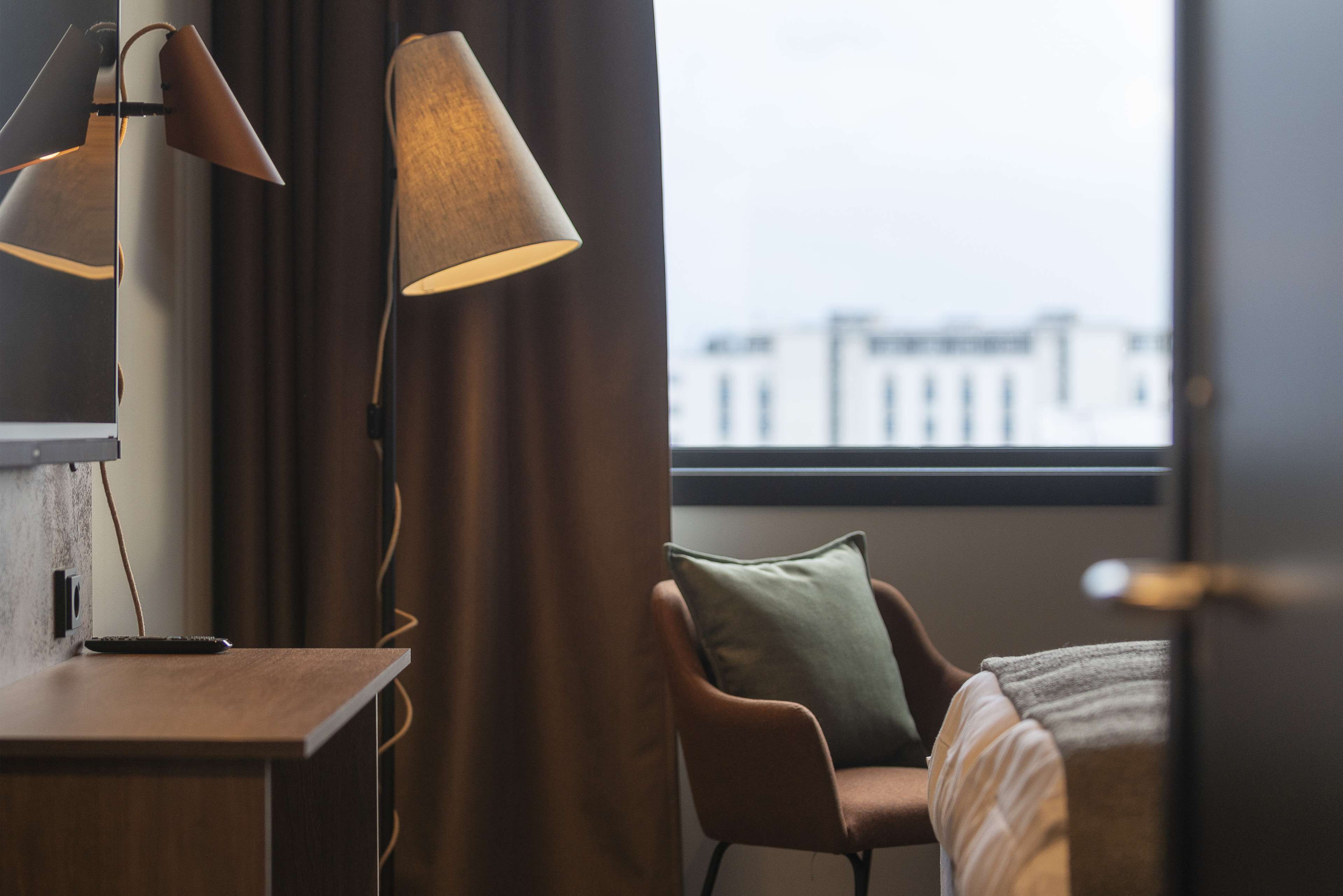 Habitación de hotel con escritorio, dos lámparas, una silla, una cama y vistas a edificios a través de una ventana.