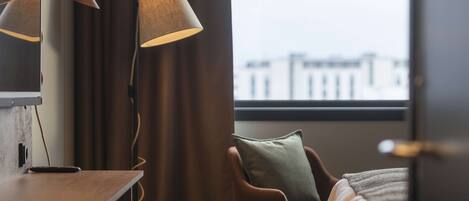 Habitación de hotel con escritorio, dos lámparas, una silla, una cama y vistas a edificios a través de una ventana.