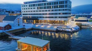 Un hôtel moderne offrant une vue sur le front de mer, avec une piscine entourée de parois de verre et un quai où est amarré un petit bateau.