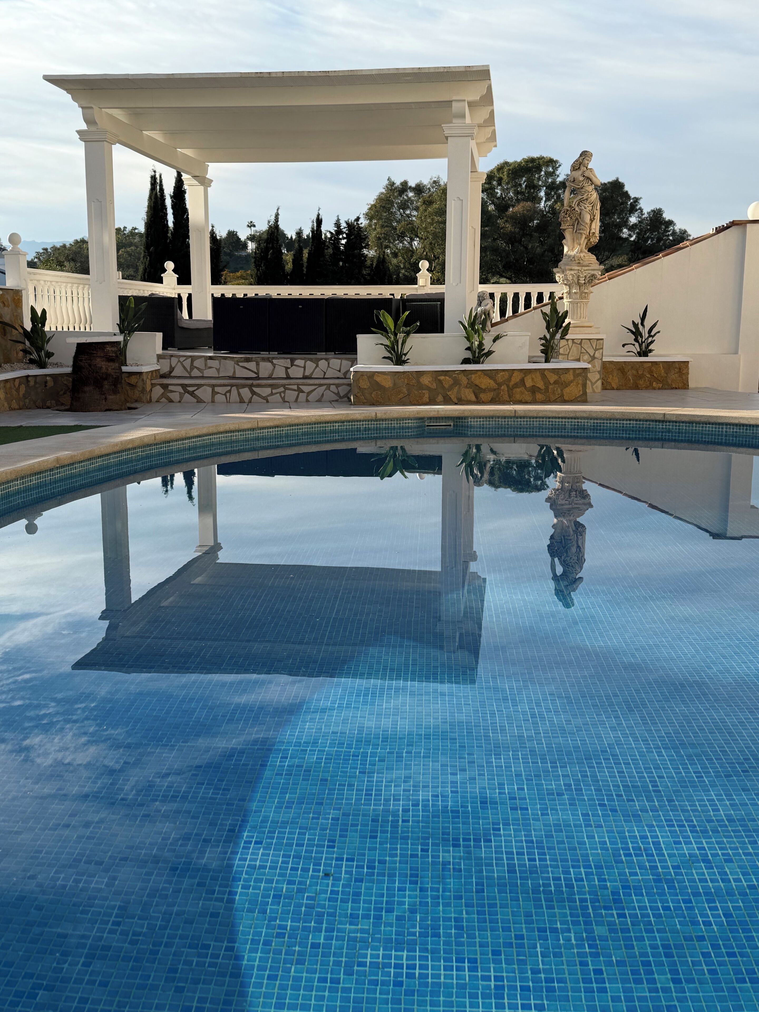 Une piscine d’eau cristalline, d’un bleu éclatant, avec une pergola blanche et une statue en arrière-plan.