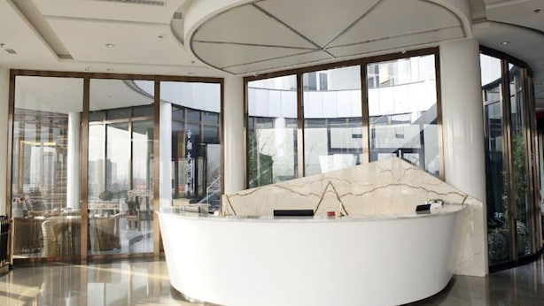 A modern reception area with a curved white reception desk, large windows offering views of the cityscape, and a glass partition separating the reception from the seating area.