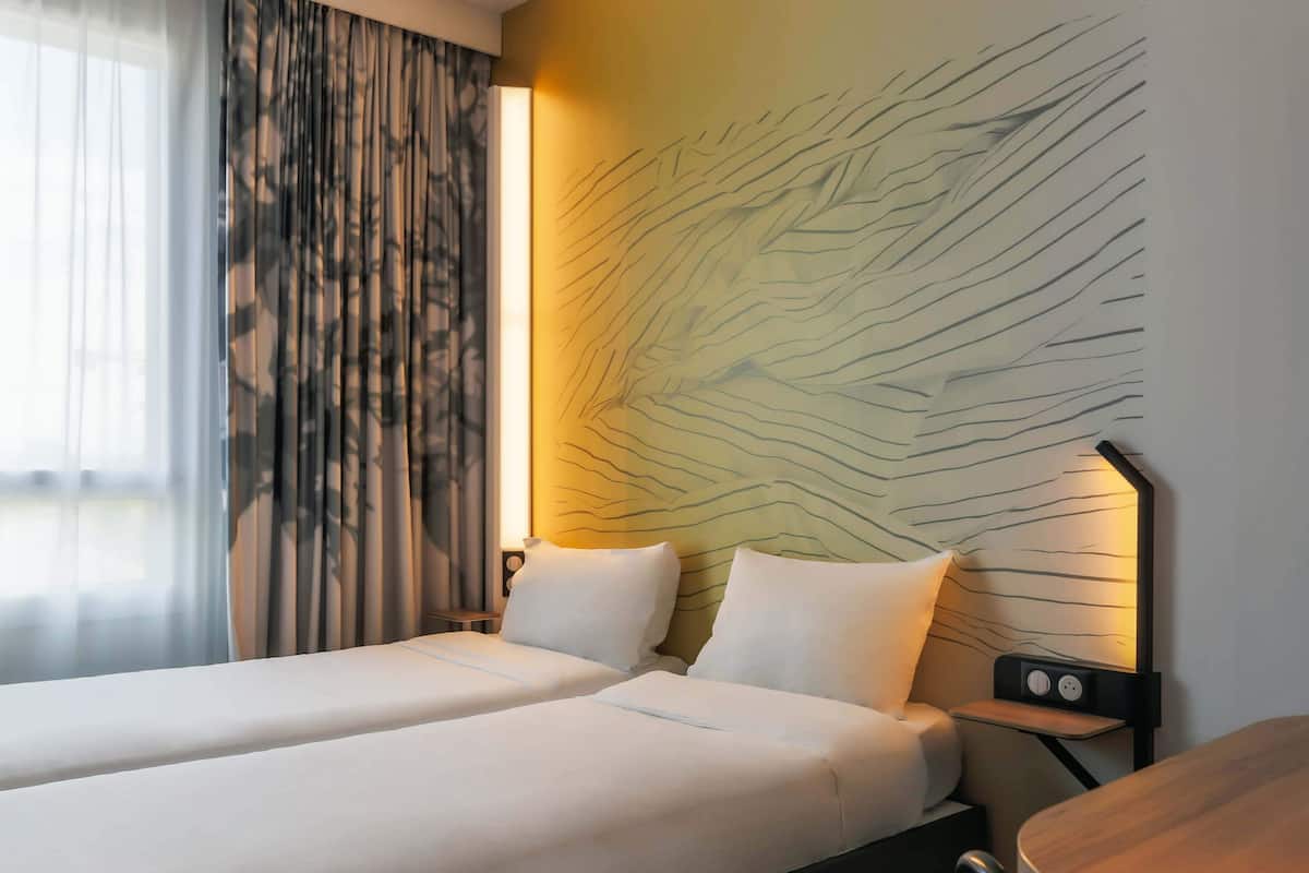 Habitación de hotel con dos camas, una mesita de noche con lámpara y una pared con diseño de hojas.