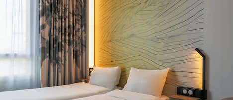 Quarto de hotel com duas camas, uma mesa de cabeceira com abajur e uma parede com padrão de folhas.