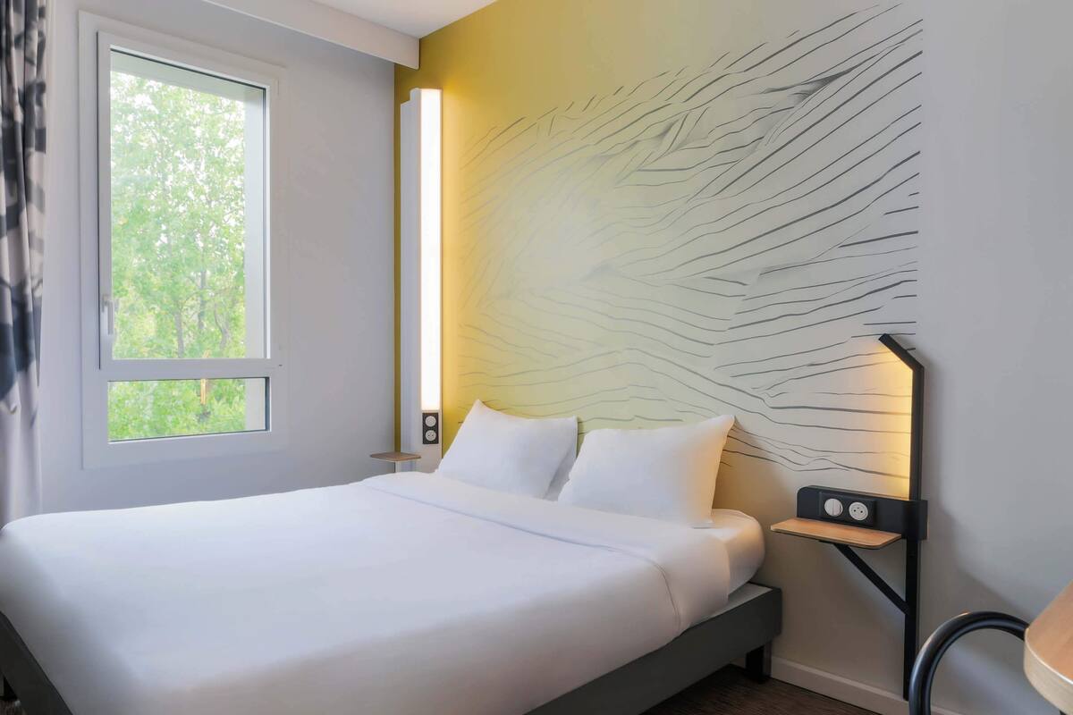 Un dormitorio moderno con cama, mesita de noche, luz de pared y ventana con vistas a árboles.