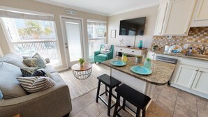 A beach house interior with a living room, kitchen, and a balcony with a view of palm trees. - Seascape 2107 1 Bedroom Condo by RedAwning (Galveston)