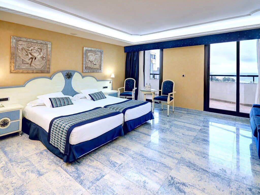A hotel room with a large bed, two chairs, a small table, and a balcony with a view.