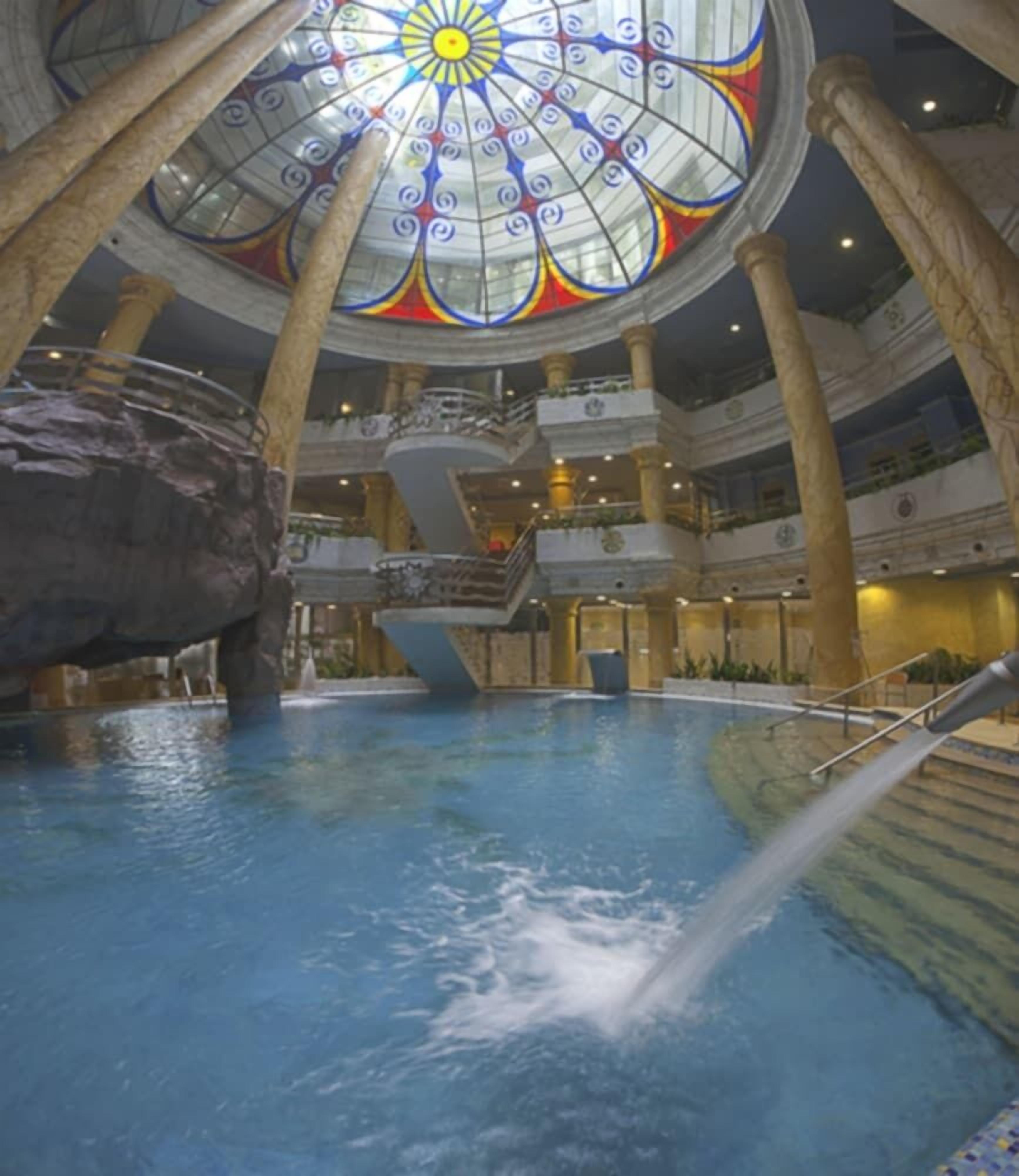 An indoor swimming pool with a central water feature, a large decorative glass ceiling, and multiple balconies.