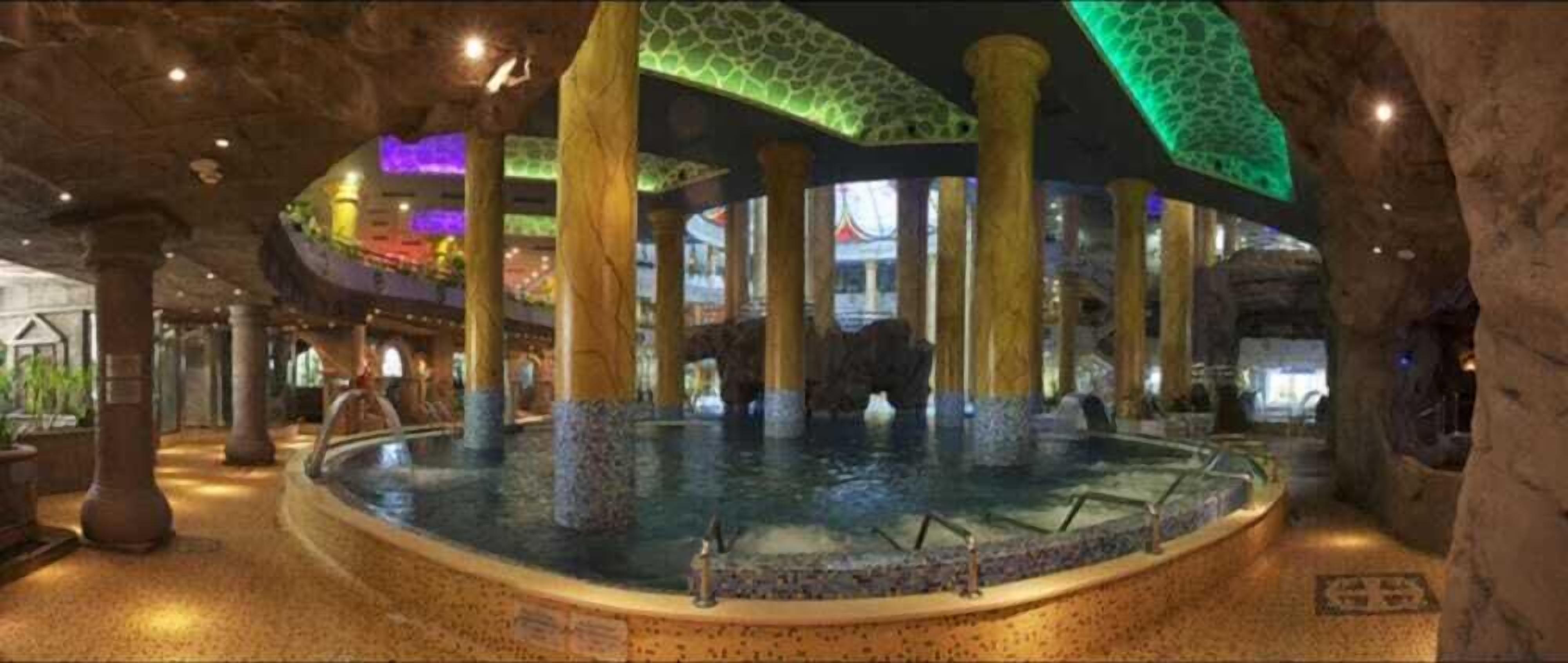 A large indoor pool with multiple columns and a central fountain, surrounded by stone walls and a tiled floor.