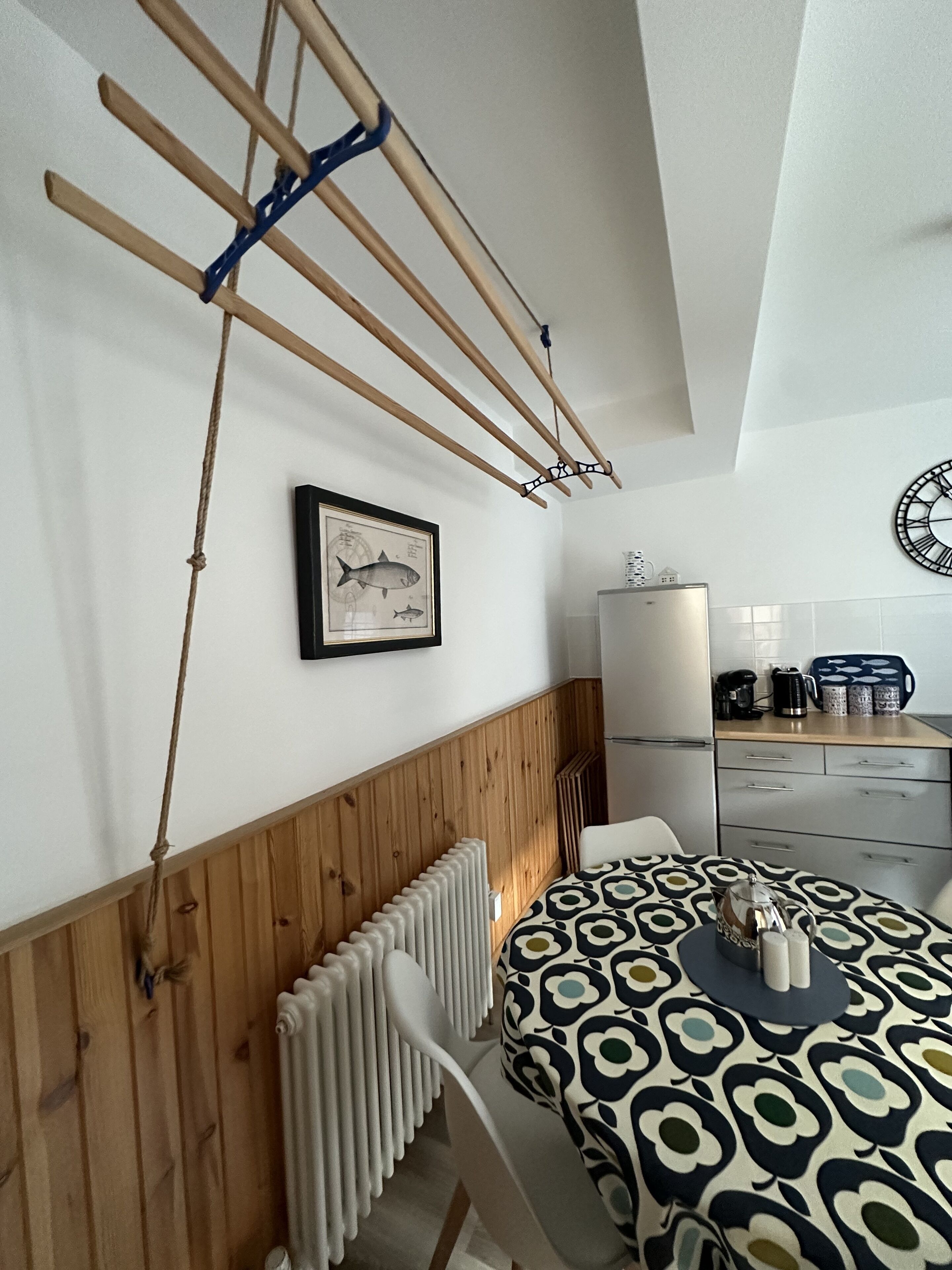 Een keuken met een houten wandpaneel, een witte radiator, een tafel met een tafelkleed met dessin, en een klok aan de muur.