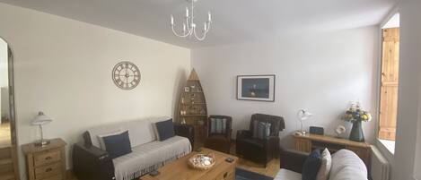 Ein Wohnzimmer mit einer Couch, einem Holztisch, einer Pendelleuchte, einer Uhr, einem Bücherregal und einem Fenster mit Blick.