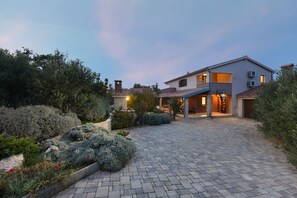 Une maison moderne à deux étages, dotée d’une allée pavée et entourée de jardins aménagés et d’arbres matures.