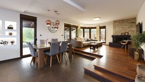 Een ruime woonkamer met een houten eettafel, grijze stoelen, een bank en een open haard.