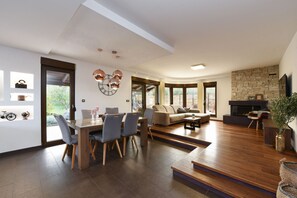 Een ruime woonkamer met een houten eettafel, grijze stoelen, een bank en een open haard.
