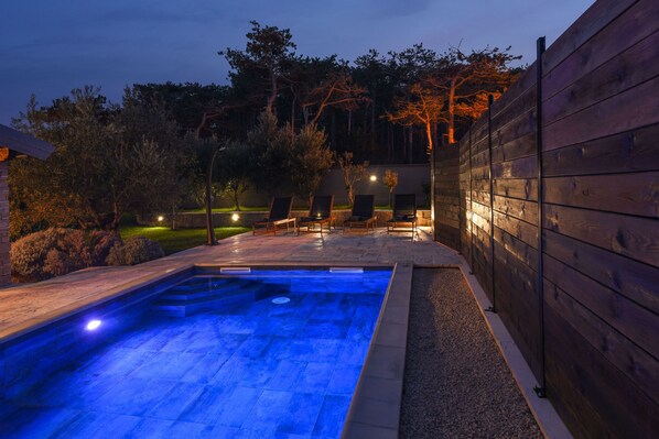 A swimming pool with a blue-lit edge, surrounded by a wooden fence, and a patio area with chairs.